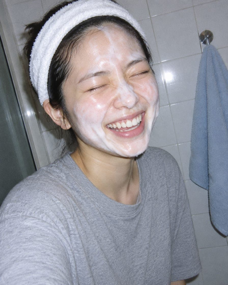 A woman in a headband laughing with cleanser foam on her face